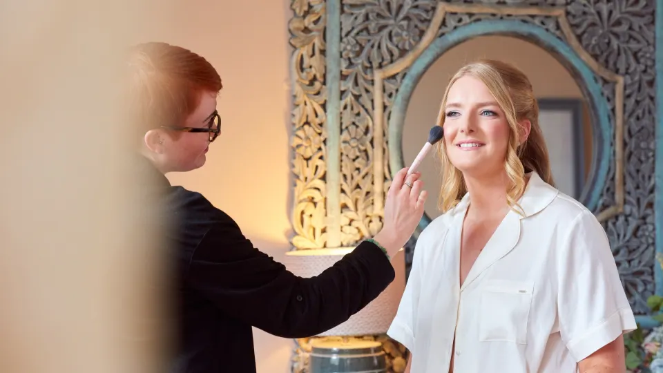 Our make-up artist sweeping blush onto a smiling bride with half-up soft waves