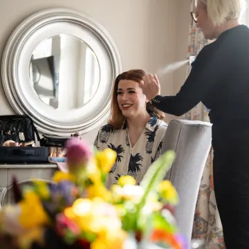 Our hairstylist setting a smiling client's red hair with hairspray at a round mirror