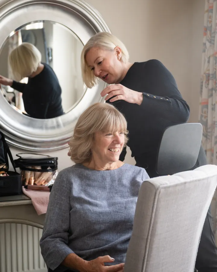 Our hairstylist styling the hair of a laughing mother of the bride at a round mirror