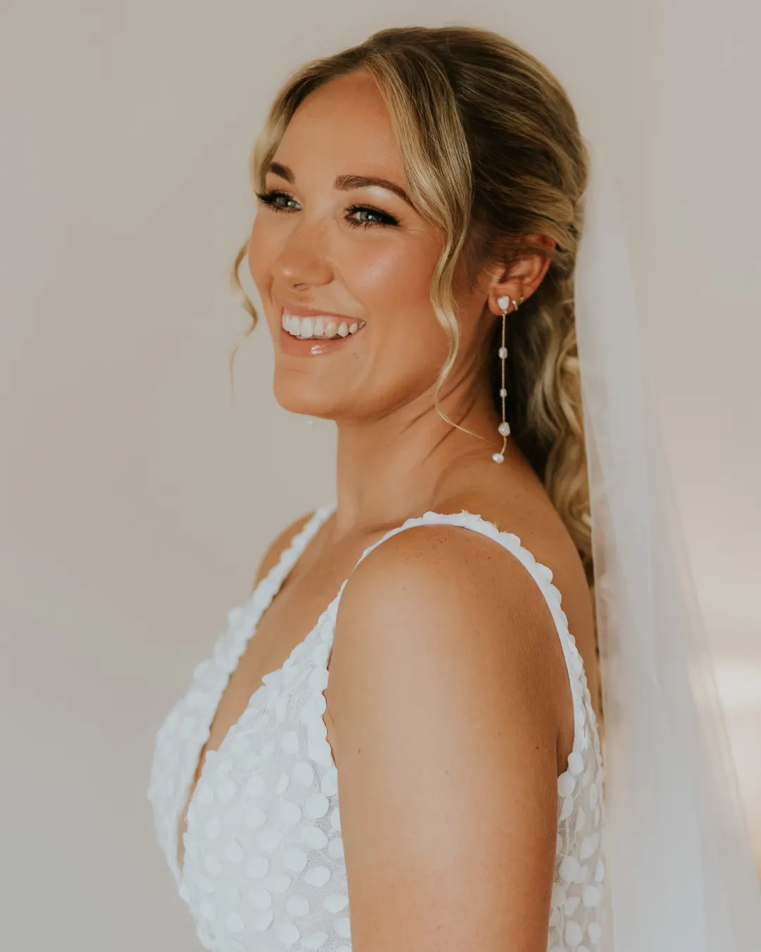 Laughing blonde bride with a half-up low ponytail of waves, curtain-bang tendrils and radiant bronzed skin