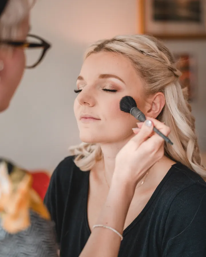 A blush brush on a bride's cheek, eyes closed, showing a bronze-gold shimmer lid and winged liner