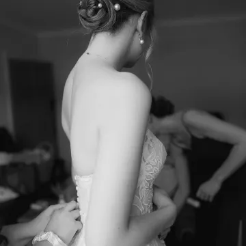 Black-and-white: a bride being buttoned into her lace gown, her braided low bun set with large pearl pins