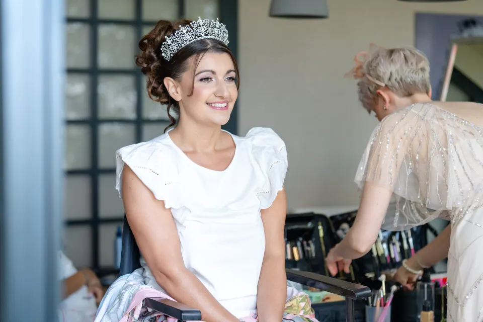 Smiling bride with a curled textured updo and a crystal tiara, soft lilac-pink eyes and a pink lip, with our make-up artist behind her
