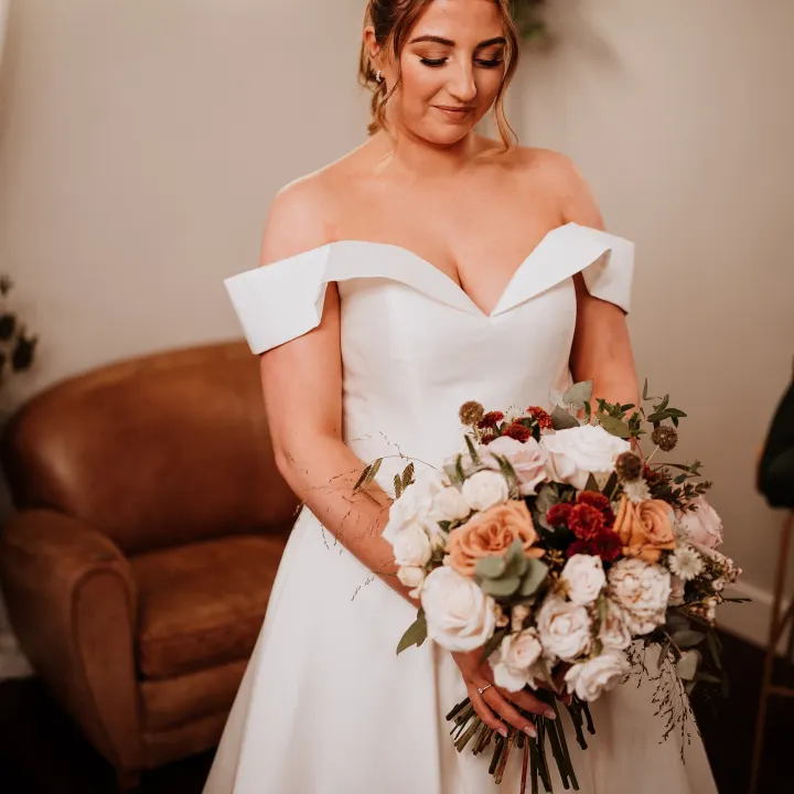 Bride in an off-shoulder satin gown looking down at her bouquet, her hair in a low bun with soft face-framing pieces