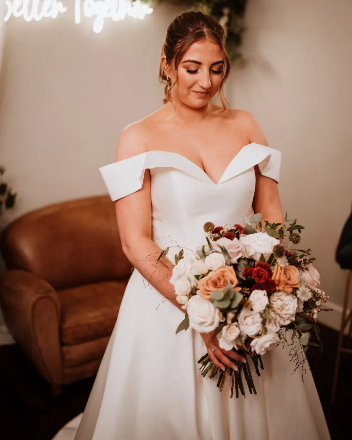 Bride in an off-shoulder satin gown looking down at her bouquet, her hair in a low bun with soft face-framing pieces