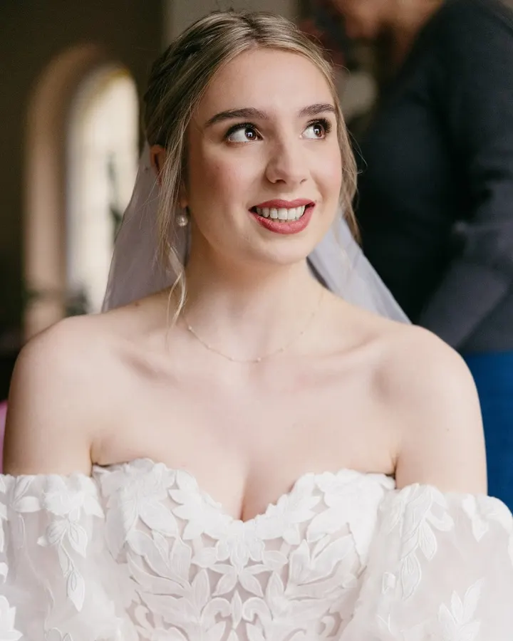 Bride smiling in an off-shoulder lace gown and veil, with soft glam and a rosy lip