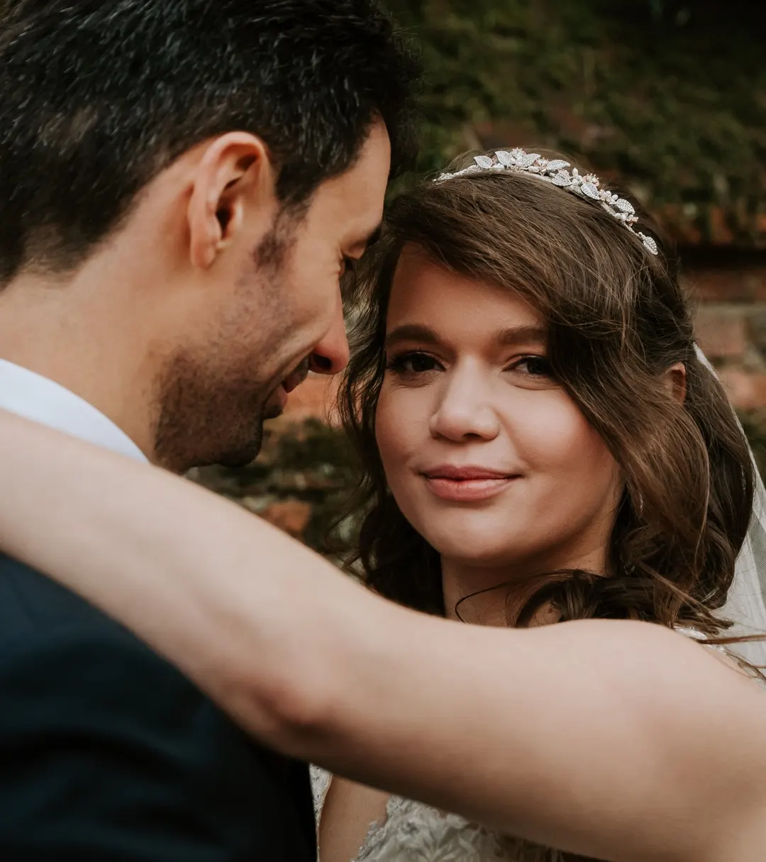 The tiara bride embracing her groom by a brick wall, with half-up curls and soft glam