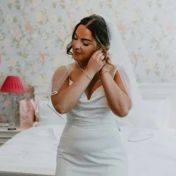 Bride putting on her earrings, with a loose low updo, face-framing waves and a veil