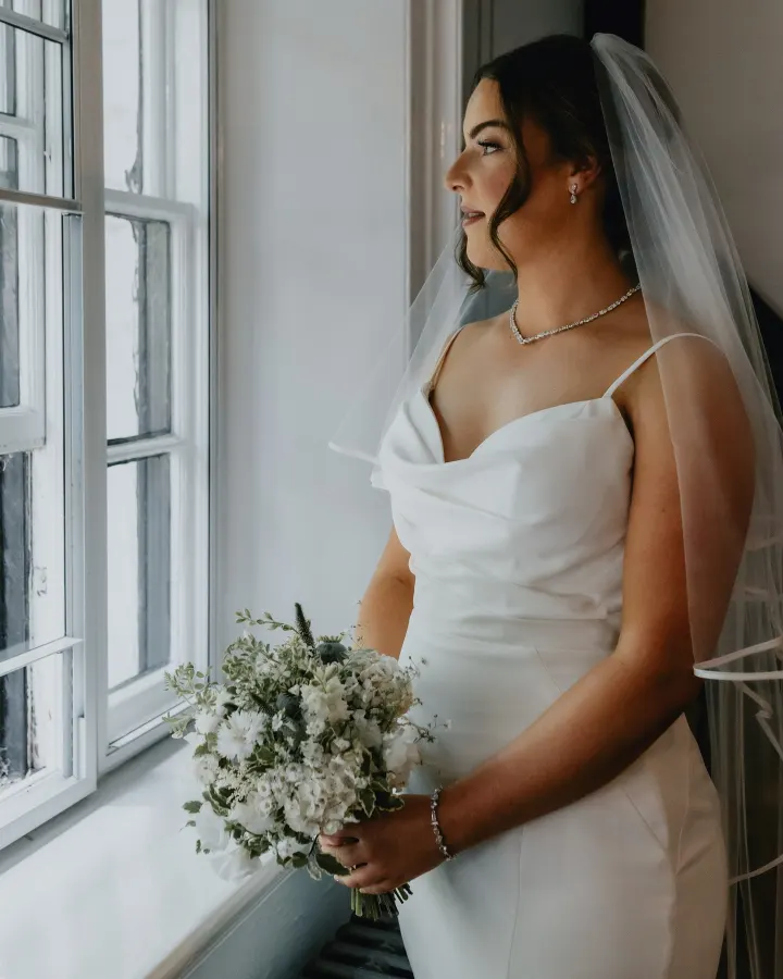 Bride in profile at a sash window: a sleek low bun with curled tendrils under a veil