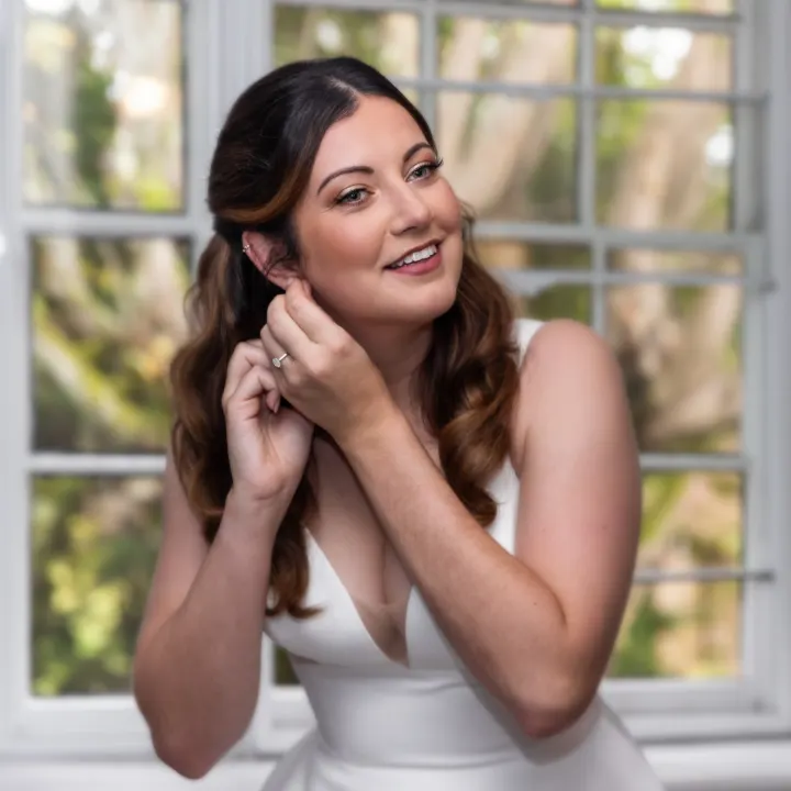 Laughing bride putting on an earring, with half-up Hollywood waves, a champagne lid and a rosy lip