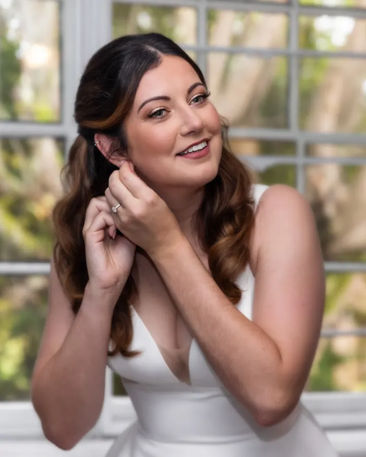 Laughing bride putting on an earring, with half-up Hollywood waves, a champagne lid and a rosy lip