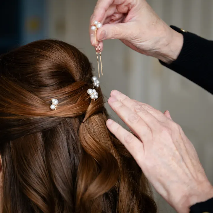 Pearl-cluster pins being placed into a crossed half-up style on glossy auburn waves
