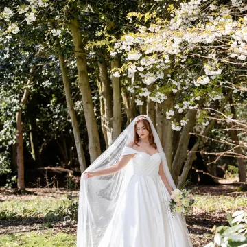 Full-length bride under blossom in a woodland clearing, holding out her veil