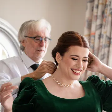 A pearl necklace being fastened for a laughing client with a sleek half-up auburn style and winged liner