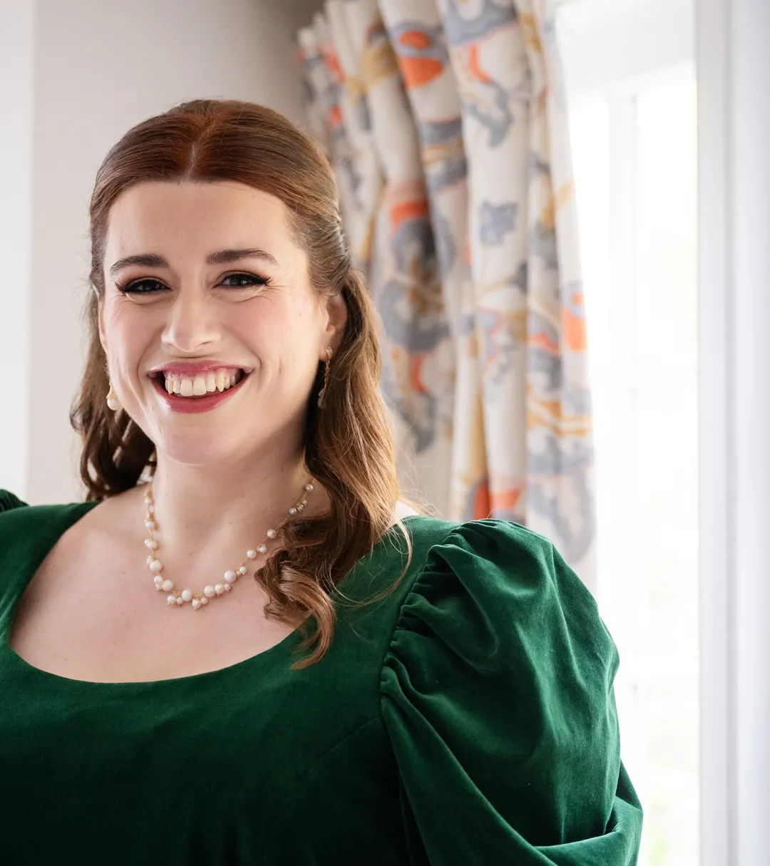 A client laughing by a sash window: half-up auburn waves, winged liner, lashes and a rose lip