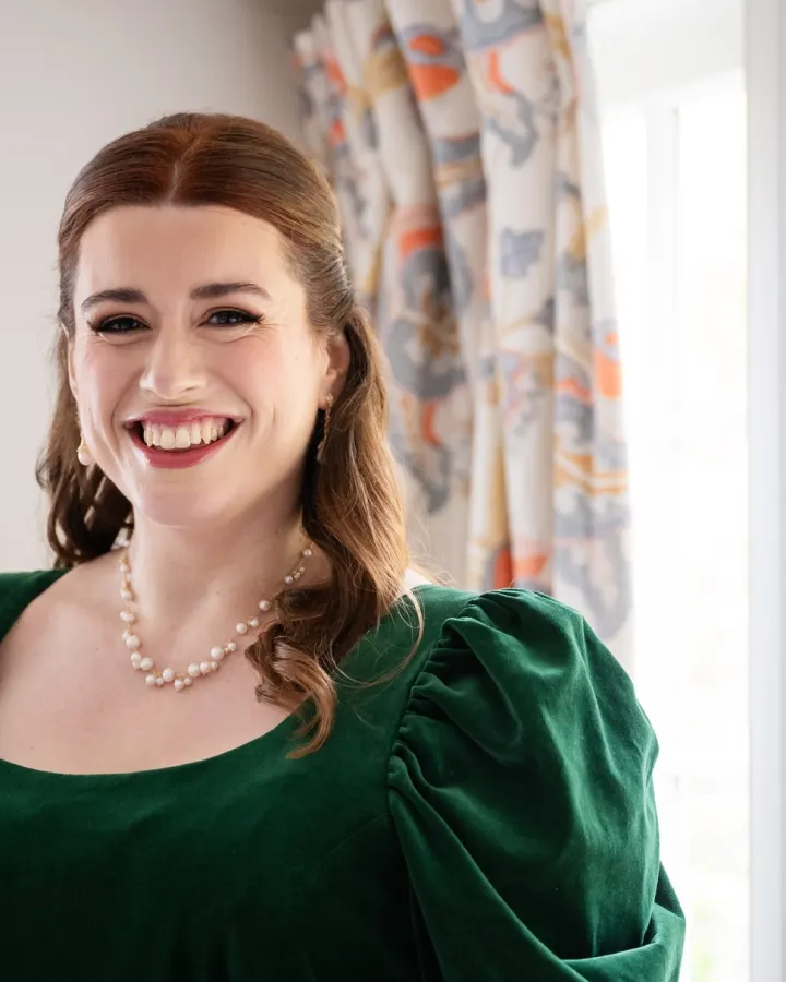 A client laughing by a sash window: half-up auburn waves, winged liner, lashes and a rose lip