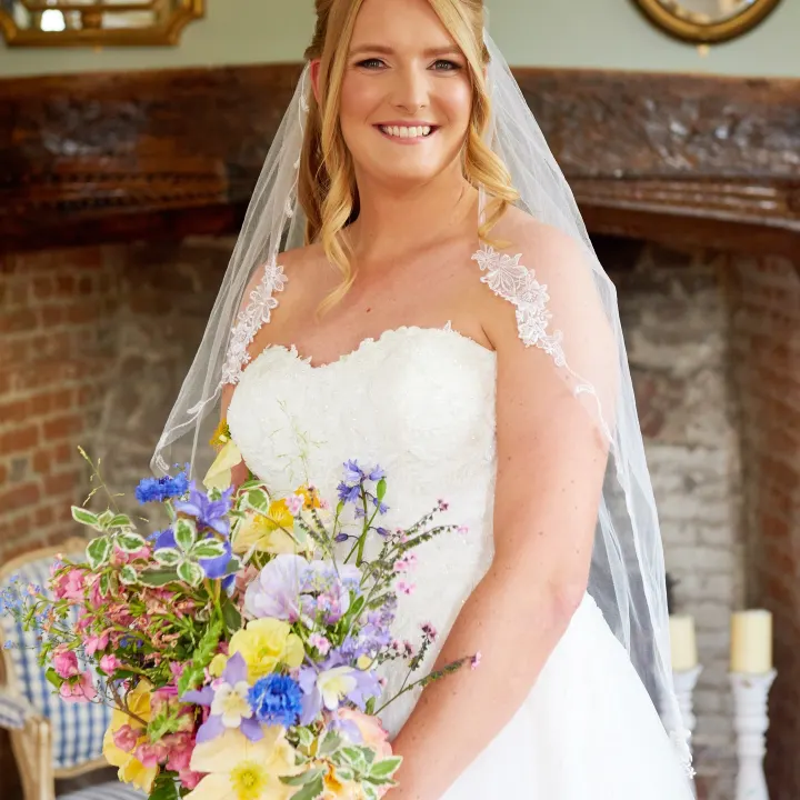Bride by a fireplace: strawberry-blonde hair half-up with soft face-framing curls, fresh glowing make-up and a pink lip