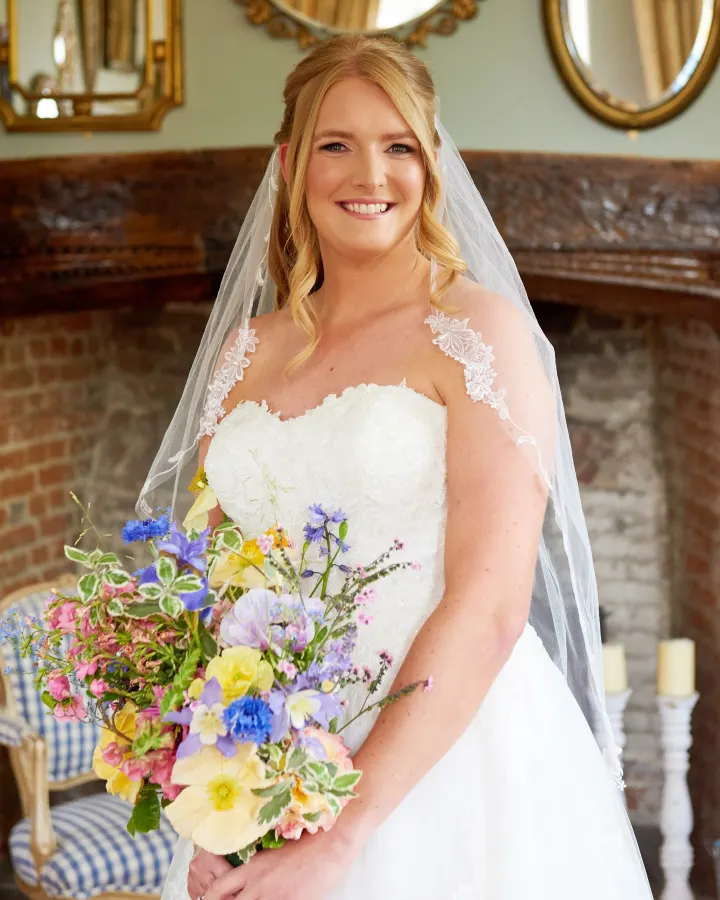 Bride by a fireplace: strawberry-blonde hair half-up with soft face-framing curls, fresh glowing make-up and a pink lip