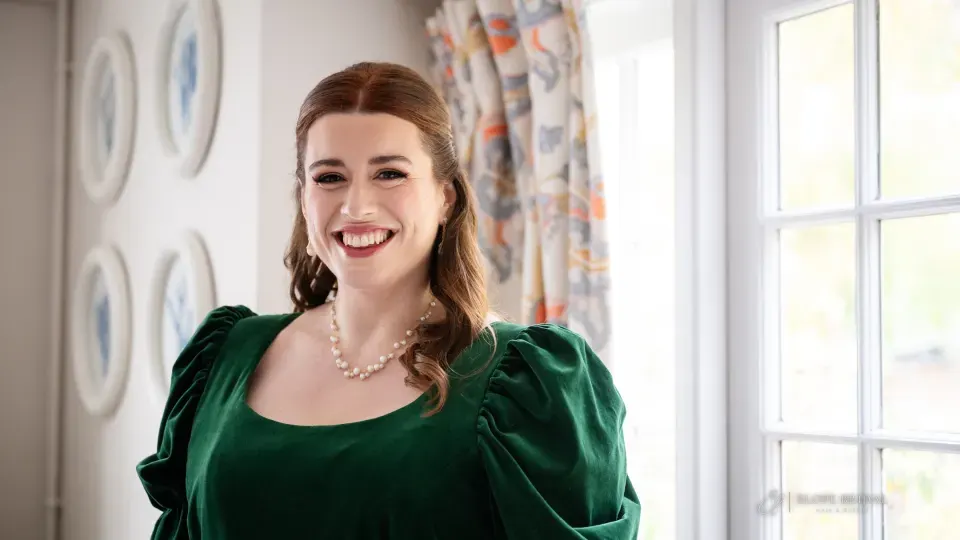 A client laughing by a sash window: half-up auburn waves, winged liner, lashes and a rose lip