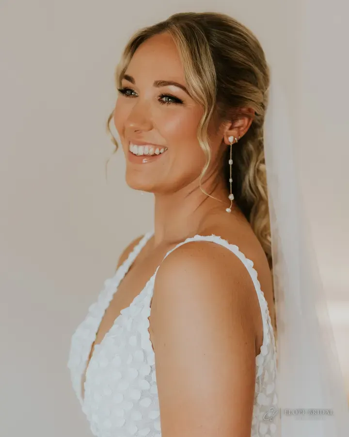 Laughing blonde bride with a half-up low ponytail of waves, curtain-bang tendrils and radiant bronzed skin